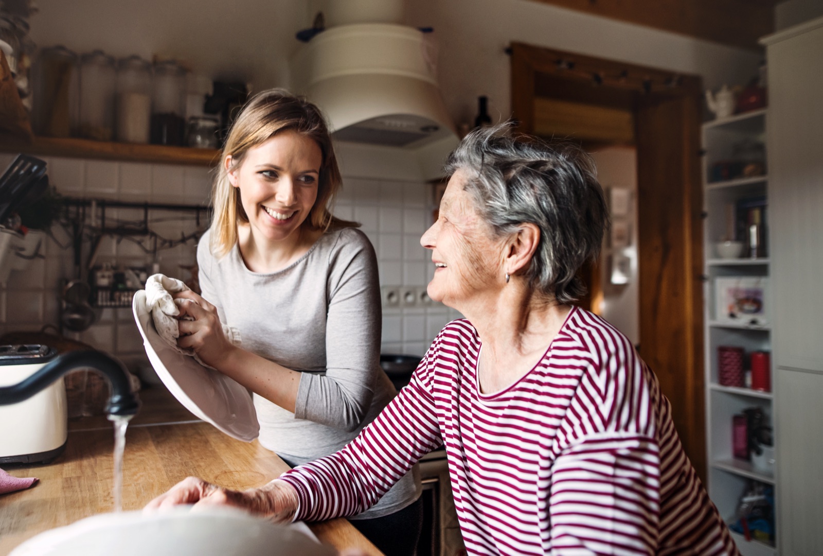 A woman and her grandmother smiling together while washing dishes in a kitchen.